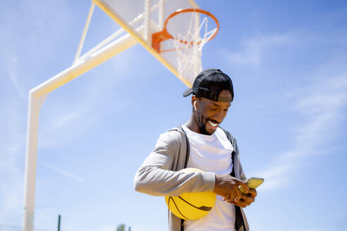 Lächelnder Mann mit Basketball und Mobiltelefon im Park - OCMF02120