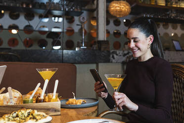 Lächelnde Frau mit Getränk, die ein Mobiltelefon benutzt, während sie im Restaurant sitzt - PNAF01730