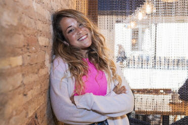 Young woman with arms crossed leaning on brick wall in bar - JRVF00886