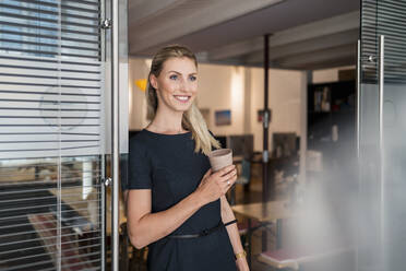 Lächelnde junge Geschäftsfrau mit wiederverwendbarem Kaffeebecher, die sich an eine Glastür lehnt und wegschaut - DIGF15303