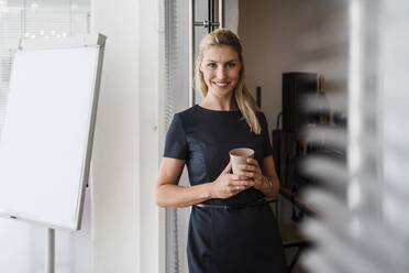 Lächelnde Unternehmerin mit wiederverwendbarem Kaffeebecher an der Tür im Büro stehend - DIGF15304