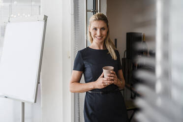 Smiling female entrepreneur with reusable coffee cup standing at doorway in office - DIGF15304