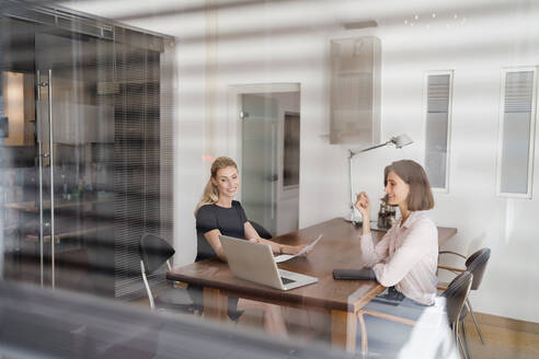 Smiling young businesswomen discussing over laptop in office seen through glass - DIGF15321