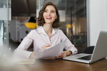 Beautiful smiling female entrepreneur sitting with document at desk while looking away - DIGF15322