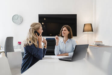 Lächelnde junge Geschäftsfrau, die eine Kollegin ansieht, die mit einem Smartphone telefoniert, während sie am Schreibtisch im Büro sitzt - DIGF15324