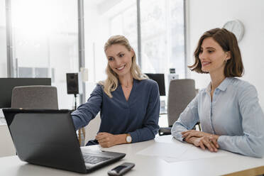 Lächelnde weibliche Kollegen schauen auf den Laptop, während sie am Schreibtisch im Büro diskutieren - DIGF15328