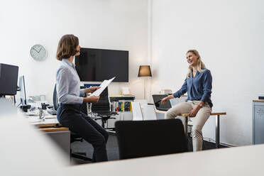 Fröhliche Geschäftsfrau im Gespräch mit einer Kollegin, während sie auf dem Schreibtisch im Büro sitzt - DIGF15329