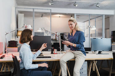 Junge Geschäftsfrau mit Blick auf eine Unternehmerin, die ein Smartphone benutzt und auf dem Schreibtisch im Büro sitzt - DIGF15334