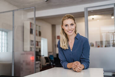 Smiling beautiful blond businesswoman leaning on desk in office - DIGF15346