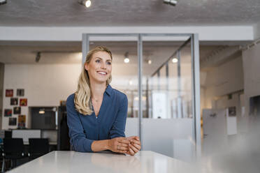 Smiling female professional looking away while leaning on desk in office - DIGF15347