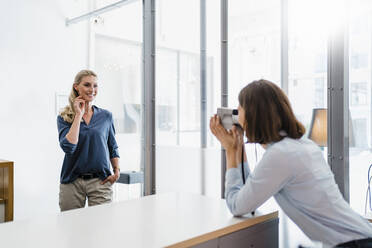 Junge Unternehmerin fotografiert eine Mitarbeiterin mit einer Sofortbildkamera im Büro - DIGF15350