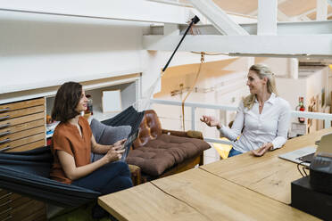 Smiling businesswoman gesturing while discussing with female professional sitting in hammock at creative office - DIGF15356