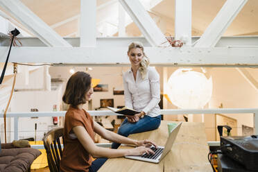 Female entrepreneur looking at colleague typing on laptop sitting at desk in creative office - DIGF15357