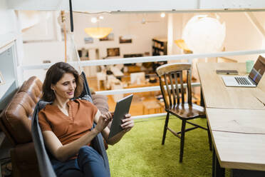 Smiling young businesswoman using digital tablet sitting in hammock at creative office - DIGF15360