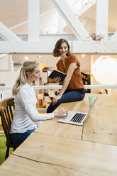 Lächelnde junge Geschäftsfrau, die auf ihren Laptop schaut, während sie mit einer Kollegin im Kreativbüro einen Plan bespricht - DIGF15363