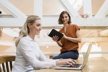 Lächelnde blonde Geschäftsfrau, die einen Laptop benutzt und neben einer Kollegin in einem kreativen Büro sitzt - DIGF15364