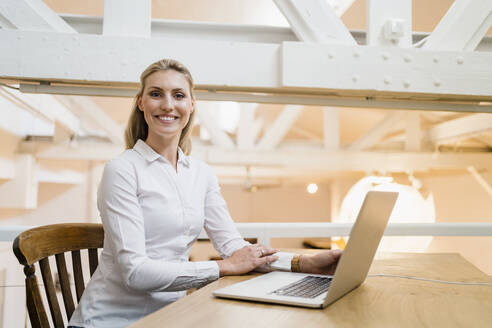 Lächelnde blonde Unternehmerin sitzt mit Laptop in einem kreativen Büro - DIGF15367