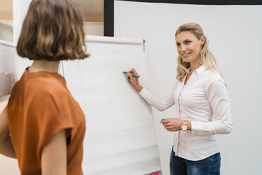 Smiling blond businesswoman writing on white board while discussing with female colleague in creative office - DIGF15371