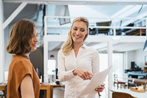 Lächelnde blonde Geschäftsfrau, die mit einer Kollegin in einem kreativen Büro über ein Dokument diskutiert - DIGF15377