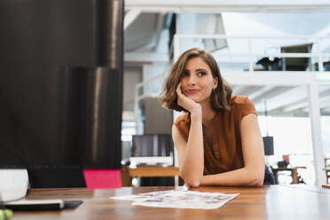 Nachdenkliche Unternehmerin, die sich in einem kreativen Büro mit der Hand am Kinn auf den Schreibtisch stützt und wegschaut - DIGF15381