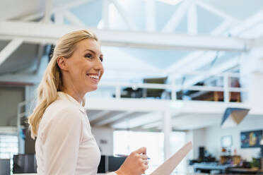 Lächelnde blonde weibliche Fachkraft mit Dokumenten in einem kreativen Büro schaut weg - DIGF15385