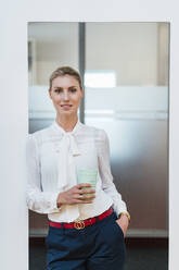 Beautiful young businesswoman standing with disposable coffee cup at doorway in office - DIGF15398