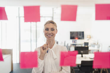 Happy female professional looking at adhesive notes seen through glass in office - DIGF15405