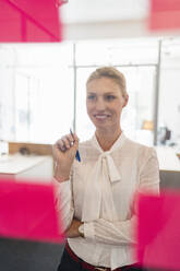 Smiling businesswoman looking at adhesive notes while working seen through glass in office - DIGF15406