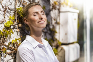 Smiling beautiful female entrepreneur standing with eyes closed in front of wall during sunny day - DIGF15413