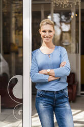 Beautiful smiling female entrepreneur standing with arms crossed at doorway - DIGF15416