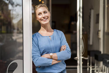 Happy businesswoman standing with arms crossed at doorway looking away - DIGF15418