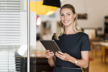Smiling blond businesswoman with digital tablet standing at doorway in office - DIGF15424