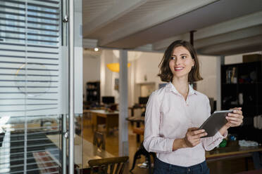 Smiling young female professional wit digital tablet standing in office while looking away - DIGF15426