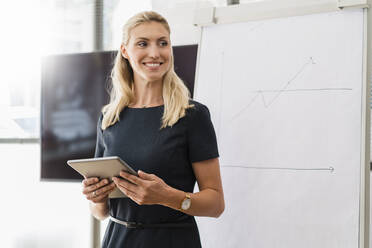 Beautiful blond businesswoman with digital tablet standing in front of white board in office while looking away - DIGF15434