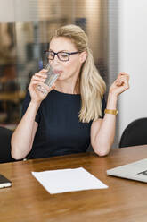 Blonde Geschäftsfrau trinkt Wasser, während sie am Schreibtisch im Büro sitzt - DIGF15437
