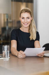 Schöne lächelnde Unternehmerin mit Dokument sitzt am Schreibtisch im Büro - DIGF15439