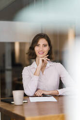 Beautiful young businesswoman sitting with hand on chin at desk in office - DIGF15449