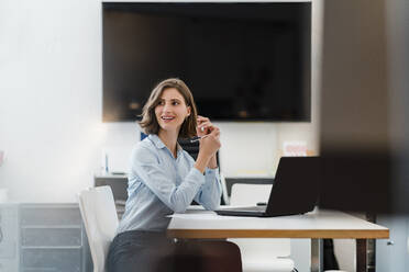 Smiling young businesswoman sitting with laptop at desk while looking away - DIGF15457