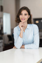 Smiling female entrepreneur with hand on chin leaning on desk while looking away - DIGF15465