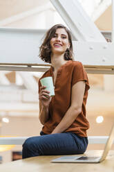 Smiling young female entrepreneur with disposable coffee cup sitting on desk at creative office - DIGF15468