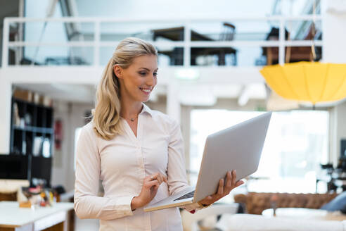 Lächelnde blonde Frau, die einen Laptop benutzt, während sie in einem kreativen Büro steht - DIGF15469