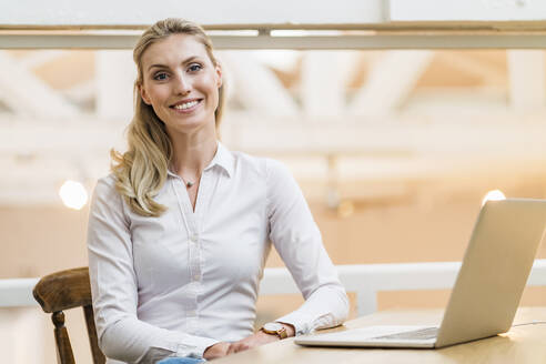 Lächelnde Unternehmerin mit Laptop am Schreibtisch in einem kreativen Büro - DIGF15471