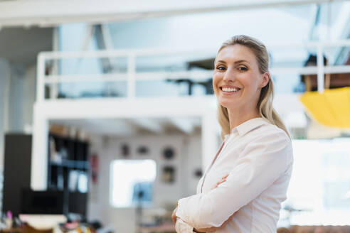 Happy blond businesswoman standing with arms crossed in creative office - DIGF15473