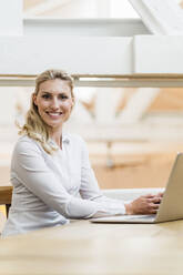 Smiling young female entrepreneur sitting with laptop in creative office - DIGF15477