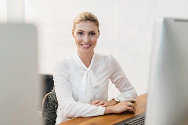 Schöne lächelnde blonde Geschäftsfrau sitzt am Schreibtisch im Büro - DIGF15482
