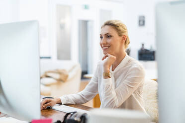 Lächelnde weibliche Fachkraft, die mit der Hand am Kinn sitzt und einen Computer im Büro benutzt - DIGF15487