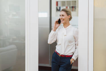 Fröhliche weibliche Fachkraft, die mit ihrem Smartphone spricht, während sie an der Tür im Büro lehnt - DIGF15490