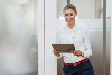 Lächelnde Unternehmerin, die ein digitales Tablet benutzt, während sie an der Tür im Büro lehnt - DIGF15492