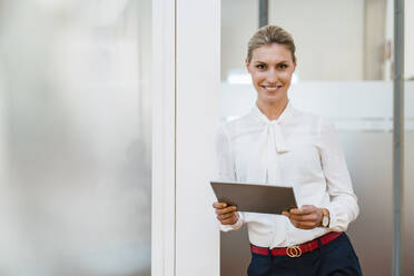 Lächelnde weibliche Fachkraft mit digitalem Tablet, die an der Tür im Büro lehnt - DIGF15493