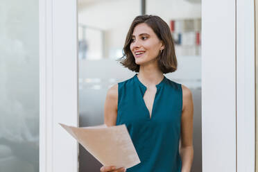 Smiling young businesswoman with documents standing at doorway in office - DIGF15494